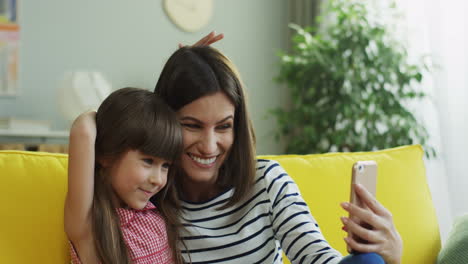 Close-up-of-the-smiled-caucasian-mother-and-daughter-taking-funny-selfie-on-the-smartphone-on-the-yellow-couch.-At-home.-Indoors.